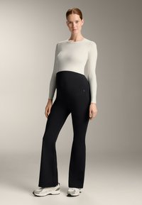 Maternity long-sleeve top in white, paired with black flared pants featuring a belly panel and pockets. Model wears gray sneakers.