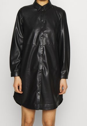 Zwarte jurk van nep leer met lange mouwen, drukknoopsluiting aan de voorkant en een puntige kraag, gedragen door een staande persoon.