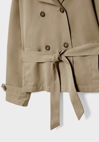 Beige double-breasted trench coat with large brown buttons, a tied waist belt, and folded cuffs. Smooth fabric with a tailored design.