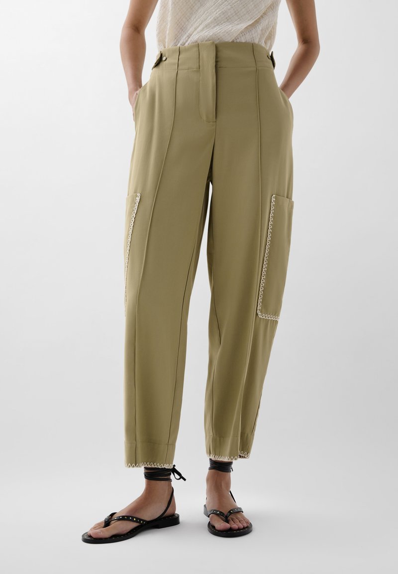 Helle olivgrüne Hose mit hoher Taille, seitlichen Taschen und dekorativer weißer Naht entlang der Nähte. Leicht zulaufendes Bein-Design.