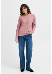 Cardigan en tricot rose avec un col rond et une fermeture à boutons sur le devant, associé à un jean large bleu et des chaussures noires. Texture lisse.