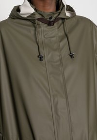Parka impermeabile verde oliva con colletto alto, fodera interna grigio chiaro a contrasto, coulisse regolabili e dettagli con chiusura a bottoni.