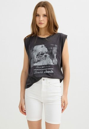 Joven mujer con cabello largo lleva una camiseta gráfica negra sin mangas que muestra un perro con gafas de sol y shorts blancos de talle alto.