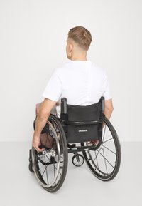 Silla de ruedas con asiento de tela negra; radios plateados y neumáticos negros; una persona ajusta individualmente la mano a la rueda trasera; camisa blanca, fondo neutro.