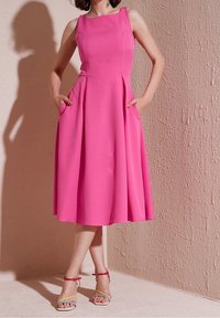 Robe sans manches rose avec un corsage ajusté, une jupe évasée et des poches latérales. Le modèle porte des sandales à lanières multicolores. Fond texturé.