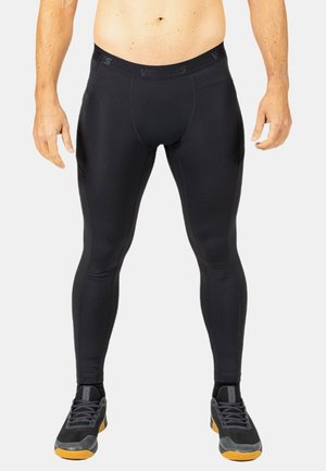 Hombre con mallas negras de compresión y zapatillas deportivas negras con suelas naranjas, de pie frente a un fondo liso.