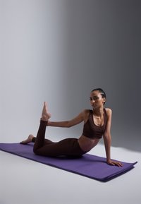 Top deportivo marrón recortado y leggings en una persona en la postura de arco sobre un tapete de yoga púrpura, contra un fondo degradado gris.