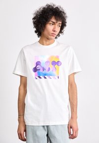 Λευκό βαμβακερό t-shirt με πολύχρωμο γραφικό σχέδιο με αφηρημένα σχήματα σε μωβ, ροζ και μπλε, και διαγώνιες ρίγες στο κάτω μέρος.