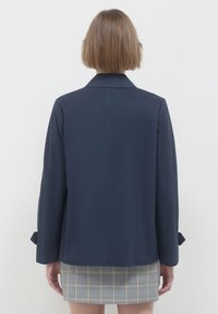 Giacca blu navy con collo, maniche lunghe e taglio dritto. Presenta dettagli ai polsini e una texture liscia. Indossata sopra una gonna a scacchi.