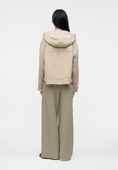 Femme aux longs cheveux noirs portant une veste à capuche beige et un pantalon large kaki, debout de dos devant un fond blanc uni.