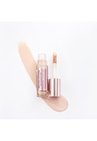 Correcteur liquide dans un tube transparent avec un couvercle en métal rose et un applicateur, à côté d'une éclaboussure crémeuse de couleur beige clair sur un fond blanc.