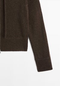 Hnědý pletený kardigan s texturovaným povrchem, který má zip a žebrované manžety. Tkanina má jemnou barevnou variaci.