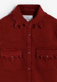Camisa de mezclilla roja con cuello puntiagudo, dos bolsillos en el pecho con solapas con bordes ondulados y botones de presión metálicos.