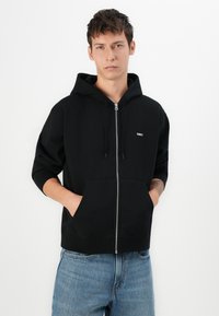 Black zip-up hoodie made of textured cotton, featuring a drawstring hood, kangaroo pockets, and a small white "OBEY" logo on the chest.