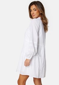 Witte katoenen jurk met lange mouwen, gelaagd ontwerp en kantaccenten op de mouwen en taille. Glad textuur, boven de knie lengte.