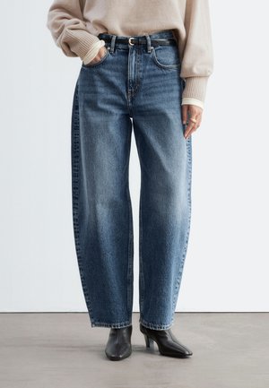 Vue recadrée d’une personne portant un jean bleu à jambes larges, un pull beige, une ceinture noire et des bottines noires. Une main est glissée dans une poche.
