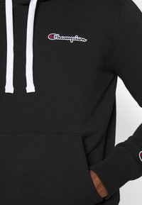 Black hoodie with white drawstrings, featuring a small embroidered logo on the chest and a pocket at the front. Smooth texture, casual design.