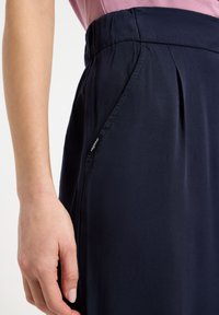 Pantaloni blu navy plissettati con vita elastica e tasca laterale; realizzati in tessuto liscio con un leggero lucido. Etichetta visibile sulla tasca.