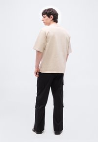 Νέος άνδρας που κοιτάζει προς τα πίσω, φορώντας μπέζ φαρδιά μπλούζα, μαύρο παντελόνι cargo και μαύρα παπούτσια, μπροστά σε απλό λευκό φόντο.