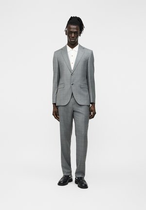 Uomo dalla pelle scura con dreadlocks, vestito con un abito grigio, camicia bianca e mocassini neri, in piedi davanti a uno sfondo bianco uniforme.