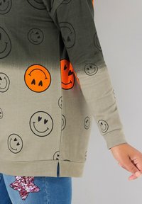 Suéter ligero de color gris-verde con estampados de caras sonrientes en negro y naranja, con una abertura lateral y tejido texturizado.