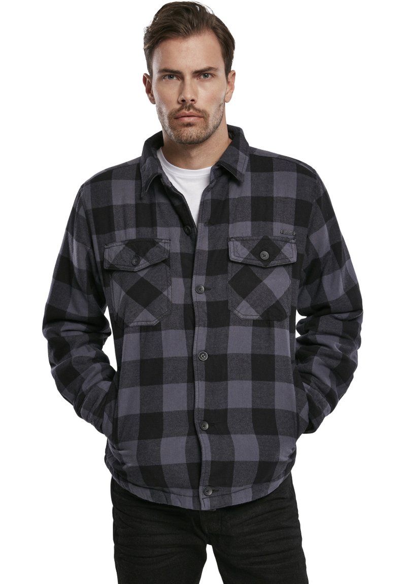 Camisa de franela a cuadros en negro y gris con dos bolsillos en el pecho, cierre de botones en la parte delantera y puños abotonados, mostrada sobre un modelo con un fondo neutro.