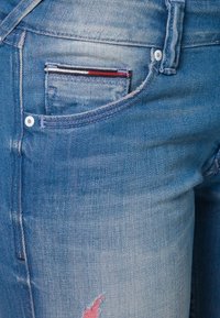 Ljusblå denimjeans med en framficka med randig fodring, en liten reva på låret och synliga sömnadsdetaljer.