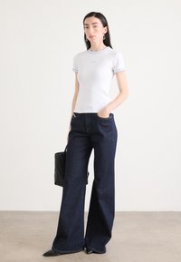 Witte t-shirt met korte mouwen en lichtblauwe afwerking, gecombineerd met donkerblauwe wijde jeans. Een zwarte handtas en spitse zwarte schoenen maken de look compleet.