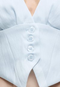 Κοντινή λήψη φωτεινού γαλάζιου υφασμάτινου ενδύματος με τέσσερα μεγάλα διαφανή κουμπιά κατά μήκος του μπροστινού μέρους και λαιμόκοψη σε σχήμα V.