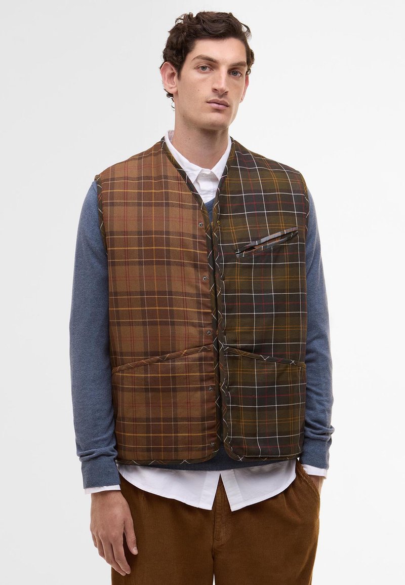 Braun und grün kariertes Weste mit Druckknöpfen, ausgestattet mit einer Brusttasche und zwei Fronttaschen. Über einem blauen Langarmshirt getragen.