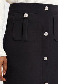 Schwarzer Rock mit tailliertem Schnitt, ausgestattet mit silbernen Knopfverschlüssen und zwei Klappentaschen. Hergestellt aus einem glatten, strukturierten Stoff.