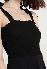 Černé šaty se zúženým korzetem, širokými ramínky a strukturální tkaninou. Obsahují mašle na ramínkách a splývání sukně.