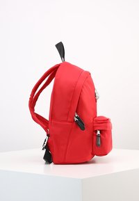 Röd ryggsäck med en slät nylonväv, med ett huvudfack och en mindre sidoficka. Dubbla axelremmar och svarta metalldetaljer.