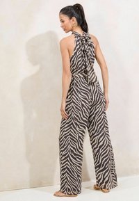 Jumpsuit uzorka zebre u smeđoj i krem boji, dizajna s halter vratom i vezanjem na leđima, širokih nogavica, lagane tkanine, i ravnih sandala.