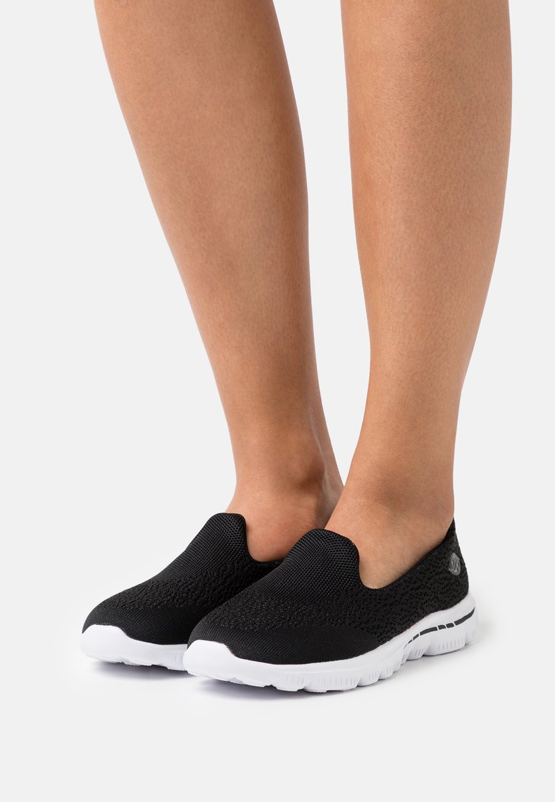 Black slip-on shoes with a textured knit upper and white rubber soles. Notable for their minimalistic design and flexible construction.