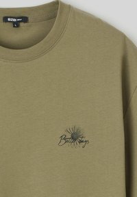 T-shirt en coton vert olive à manches courtes, col rond côtelé, et motif "Meilleurs Jours" en encre noire avec un design de pissenlit.