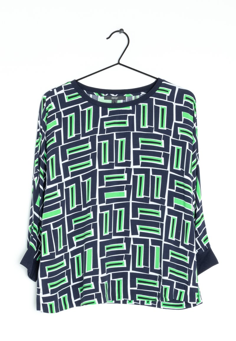 Chemise à manches longues avec un motif géométrique rectangulaire bleu marine, blanc et vert, suspendue sur un cintre en fil noir sur fond blanc.