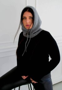 Gray knit hood with drawstrings, ribbed texture, worn over a black long-sleeve top; seated pose with smooth black leggings.