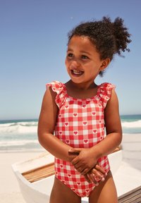Roter und weiß kariert Badeanzug mit Herzmustern, Rüschen an den Trägern und enger Passform. Ein Kind steht am Strand, Sand an den Armen.