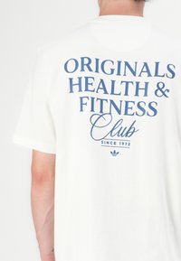T-shirt en coton blanc à manches courtes, avec le texte bleu "ORIGINALS HEALTH & FITNESS Club" et "SINCE 1972" imprimé au dos.