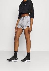 Musta lyhyt takki, jossa on läpinäkyvä huppu, harmaat tuktukkishortsit, mustat sukat ja mustat Nike-kengät valkoisilla yksityiskohdilla.
