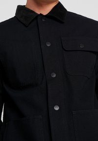 Zwarte button-up jas met een corduroy kraag, twee borstzakken en een gladde stoftextuur. Zwarte knopen voltooien het ontwerp.