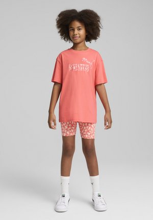 Camiseta de algodón color coral con el logotipo blanco de "PUMA", combinada con pantalones cortos rosa con un estampado ligero, calcetines de caña corta blancos y zapatillas blancas.