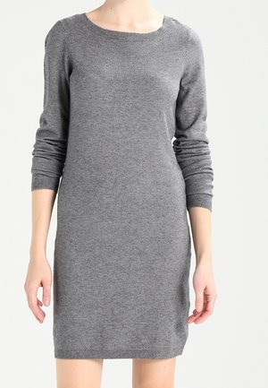 Kvinde iført en tætsiddende, langærmet, knælængde grå strikkjole, stående med armene afslappede.