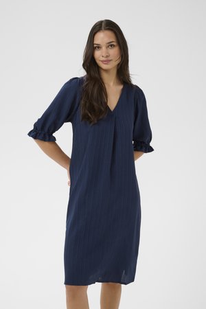 Hosszú sötét hajú nő, térdig érő tengerészkék ruhában, melynek puffos, háromnegyedes ujja és V-nyakkivágása van, kezét a hát mögött összekulcsolva áll.