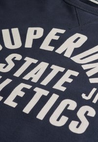 Marineblauwe sweatshirt met vetgedrukte witte tekst "SUPERDRY STATE ATHLETICS." Ronde halslijn met stikdetails, zachte textuur.