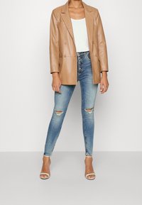 Brun läder dubbelknäppt blazer över en vit tanktop. Åtsittande blå skinny jeans med slitna knän, kombinerat med vita klacksandaler.