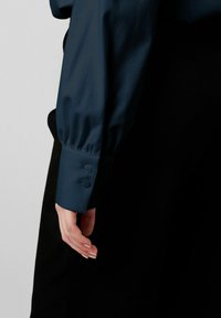 Dunkelblaues Blusenshirt mit langen Puffärmeln und Knopfmanschetten, neben einem schwarzen Kleidungsstück präsentiert, das die glatte Textur des Stoffes betont.