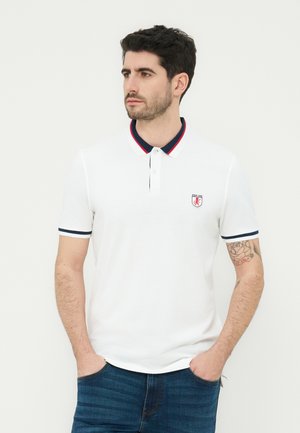 Homme aux cheveux courts et foncés portant un polo blanc avec des détails de col rouges et bleu marine, ainsi qu'un jean bleu, debout les mains dans les poches.