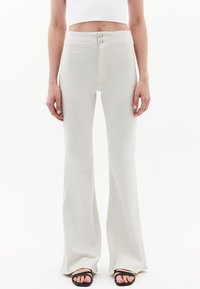 Höga, utsvängda byxor i ljusbeige tyg. Har två framknappar, sidfickor och en jämn textur. Åtföljs av svarta sandaler.
